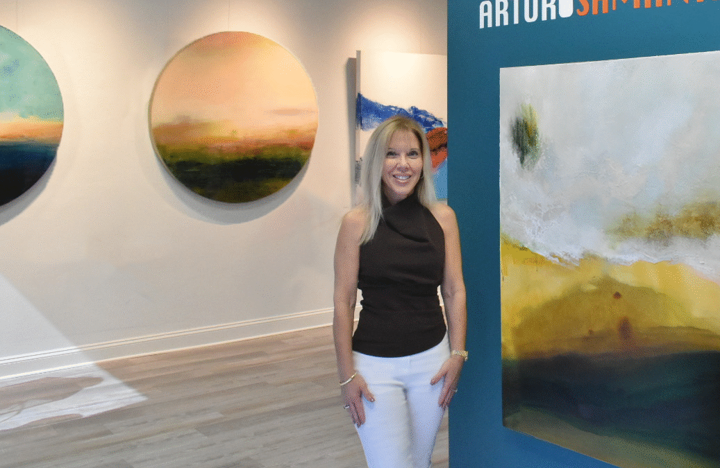 A woman stands beside abstract paintings in an art gallery.